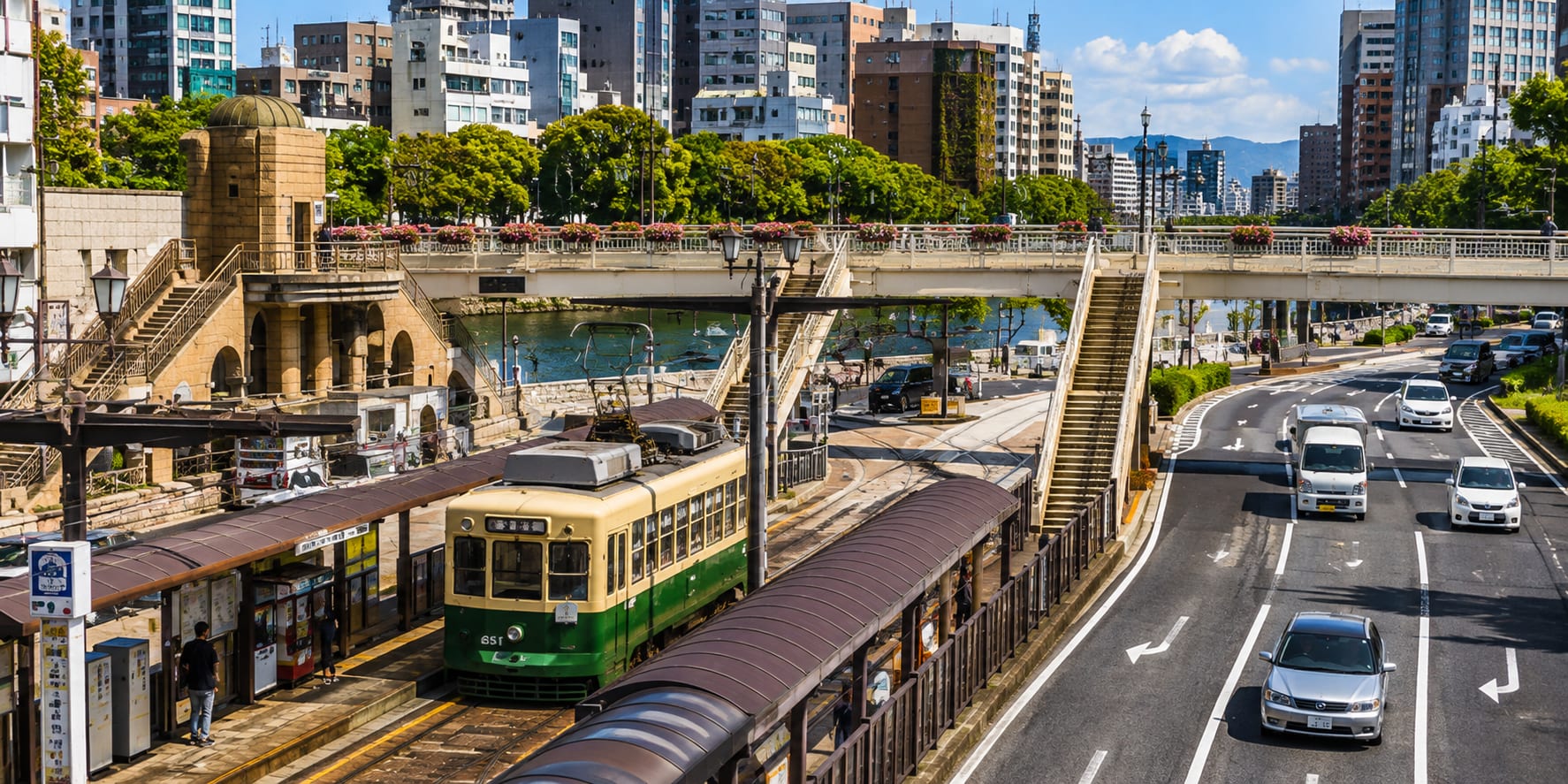 Hiroshima streetcar at a station beside the river, with city buildings and a bridge in the background
